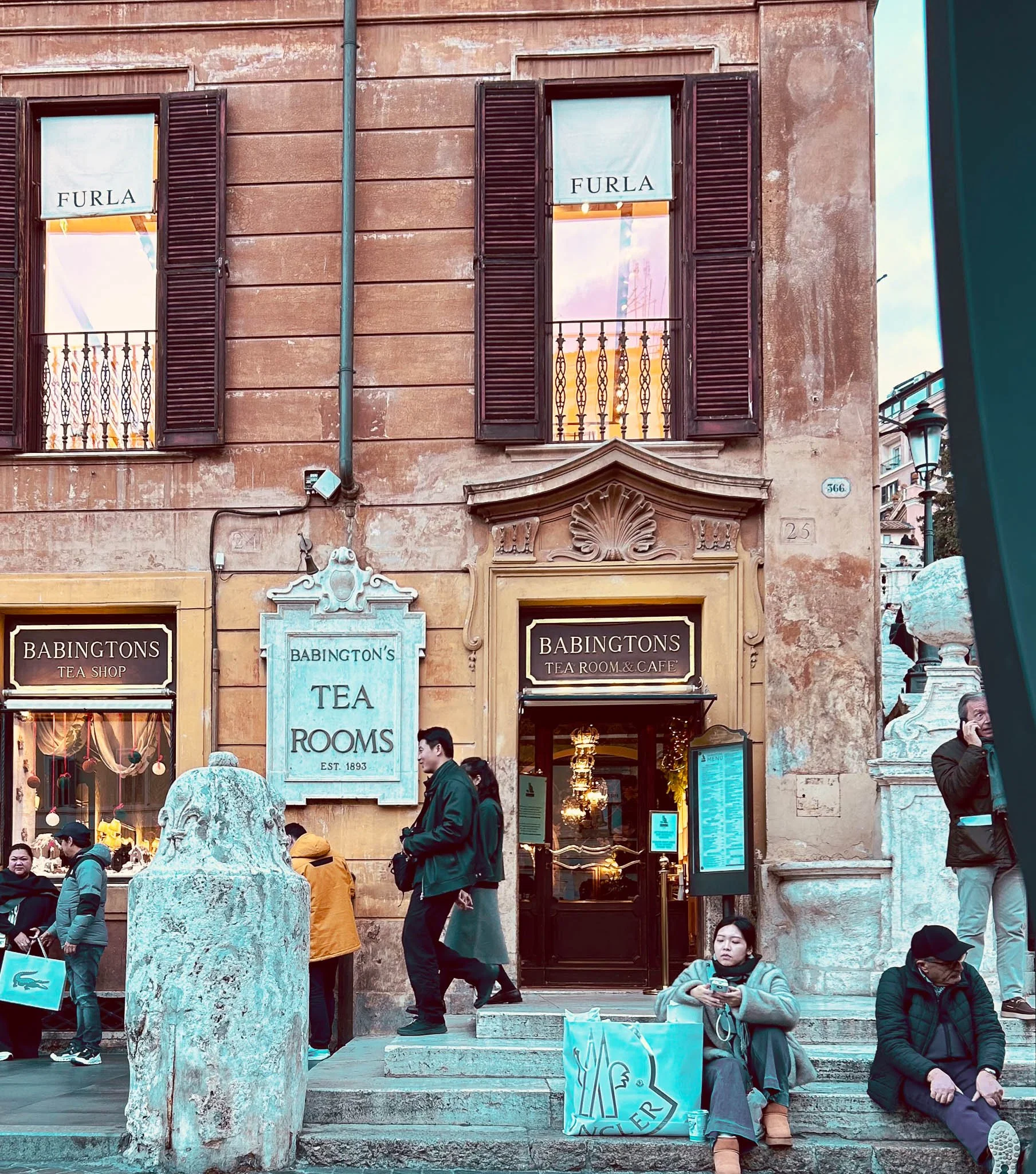 People sitting and standing outside Babington's Tea Room & Cafe, a historic tea shop established in 1893, on a busy street with various storefronts and a stone sculpture in front.