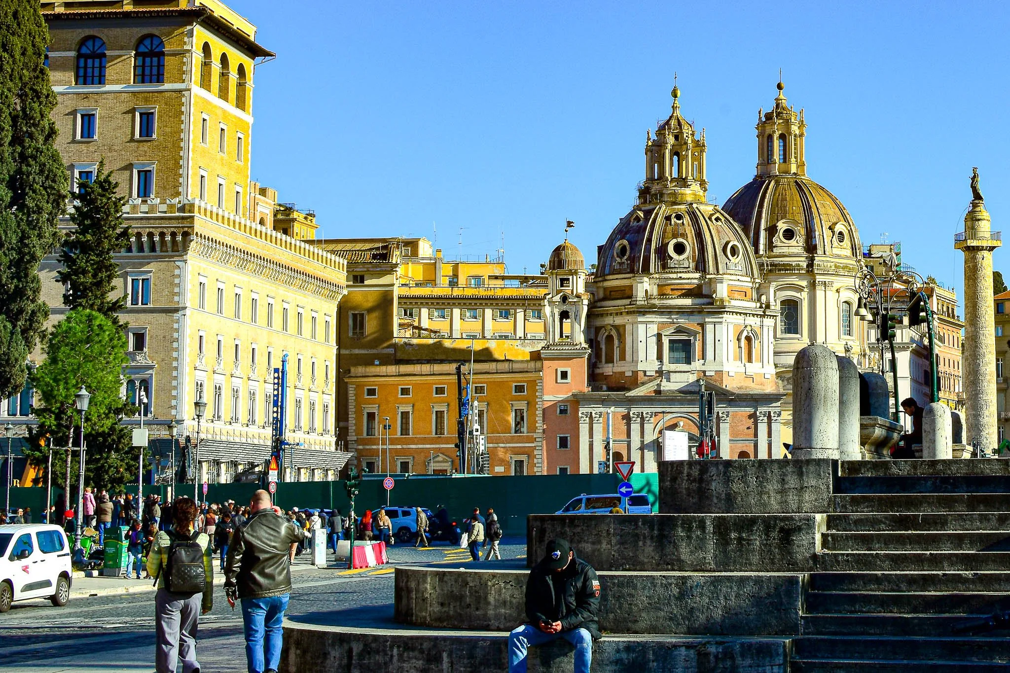 City square with historic buildings, including a church with domed towers, and people walking and sitting on stone steps. Bright and sunny day.
