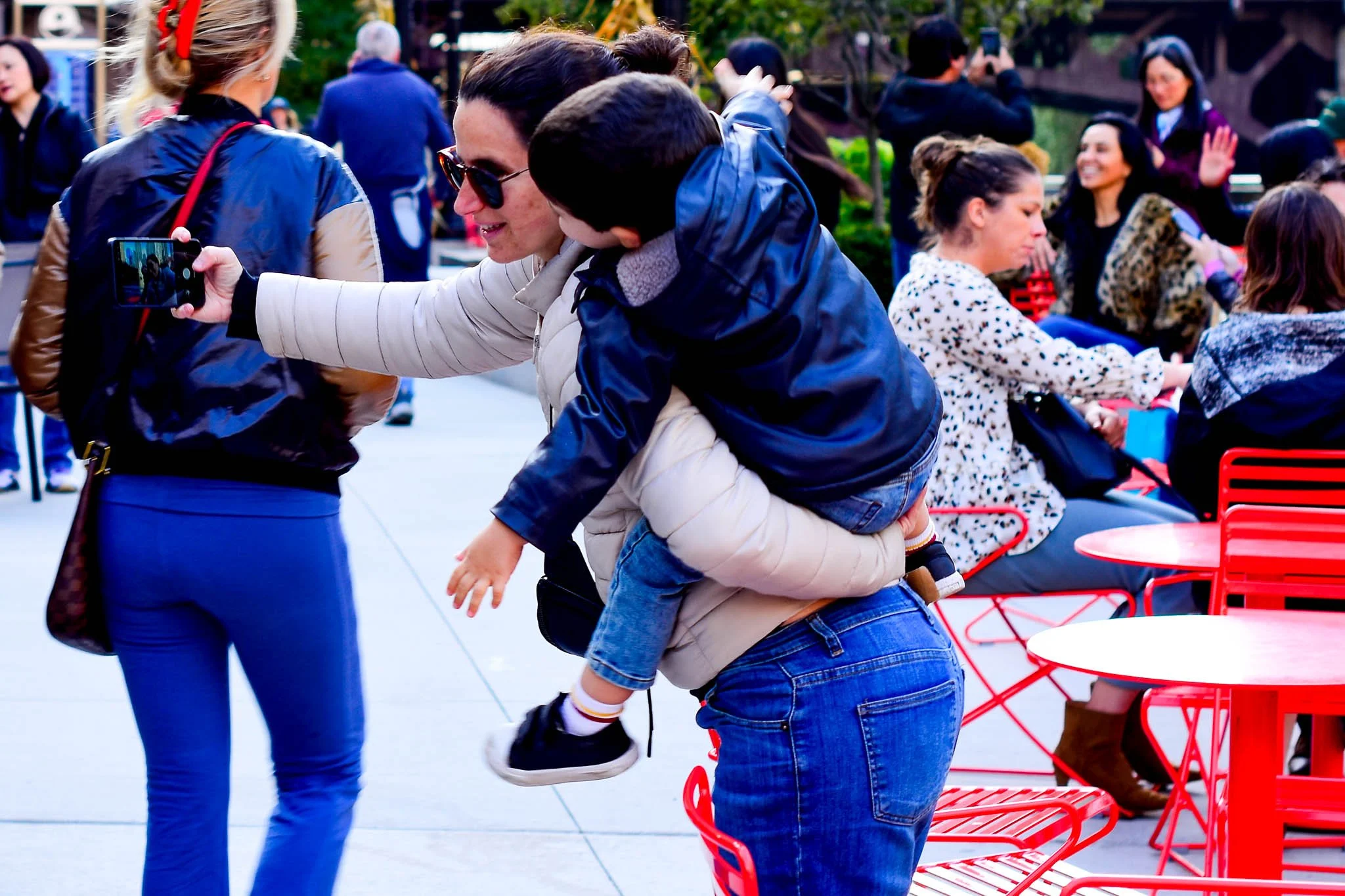 A man carrying a child on his back while taking a selfie with a woman at an outdoor social gathering, with other seated and standing people in the background.
