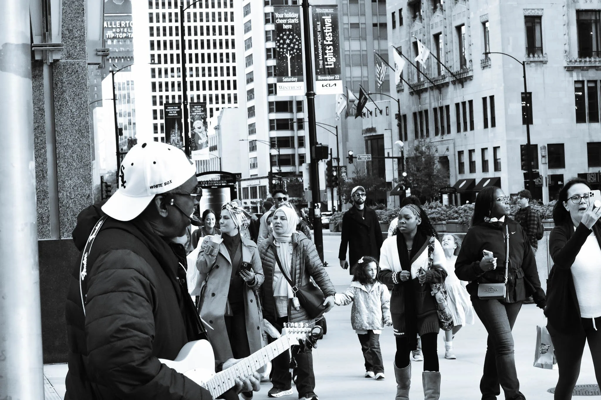 A street scene with a man playing a guitar and singing, surrounded by pedestrians in an urban area with tall buildings and street signs.