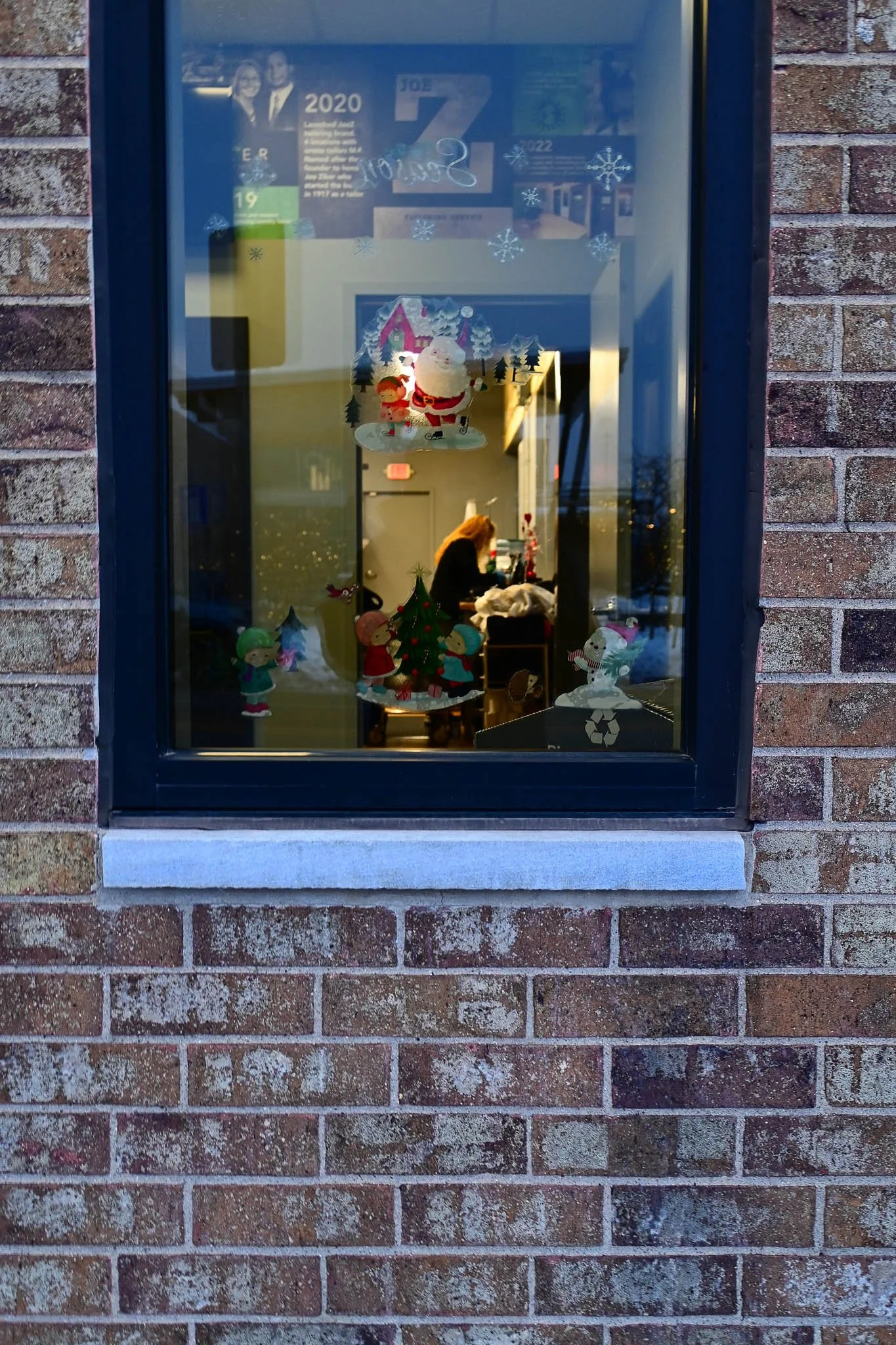 Window display with Christmas decorations, including paper cutouts of children and Santa Claus, and a person decorating inside the building.