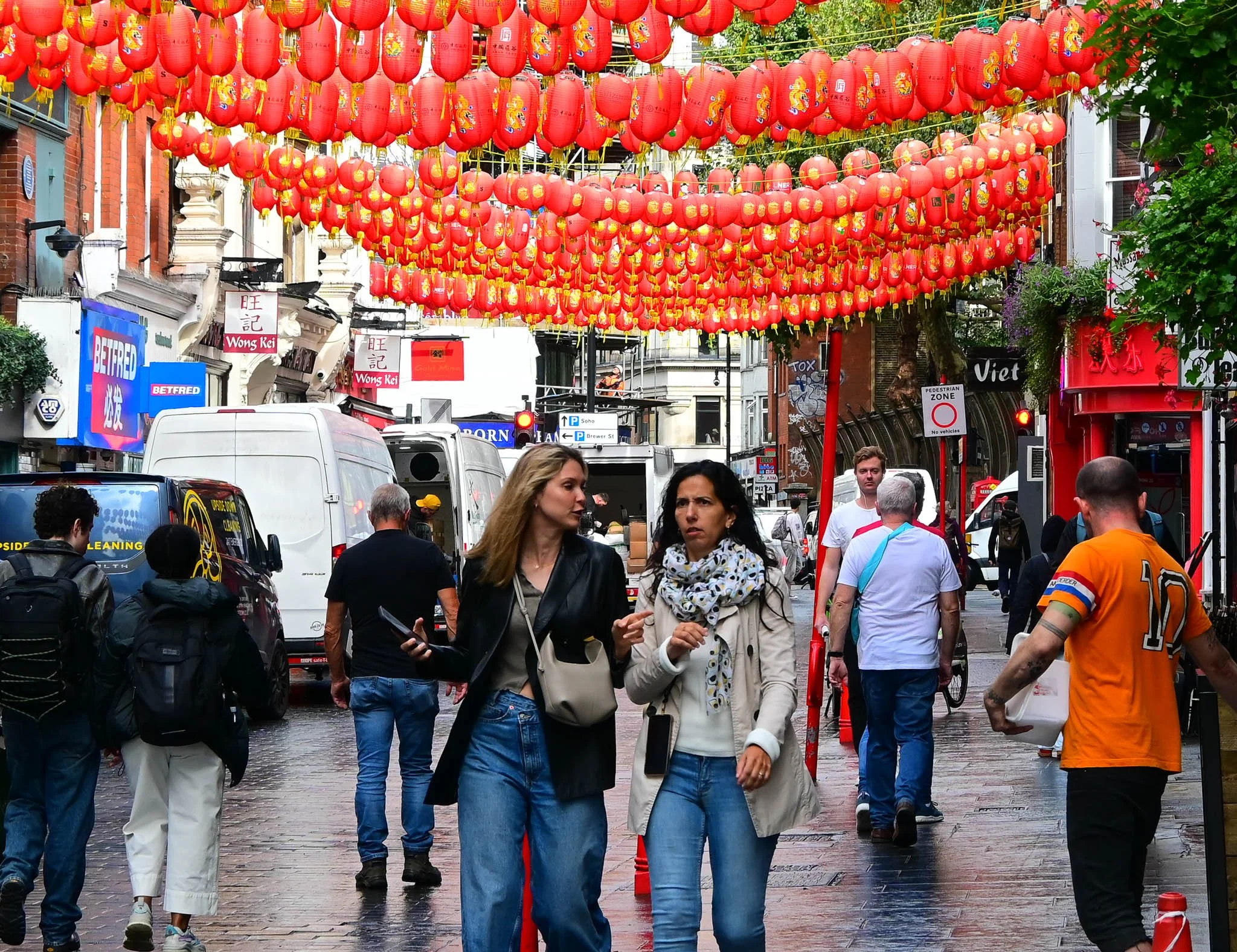Street scene decorated with red Chinese lanterns hanging overhead, with pedestrians walking on a wet pavement and various storefronts in the background.