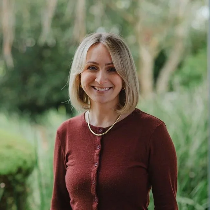 A lady in her twenties wearing an orange shirt in front of bushes outside