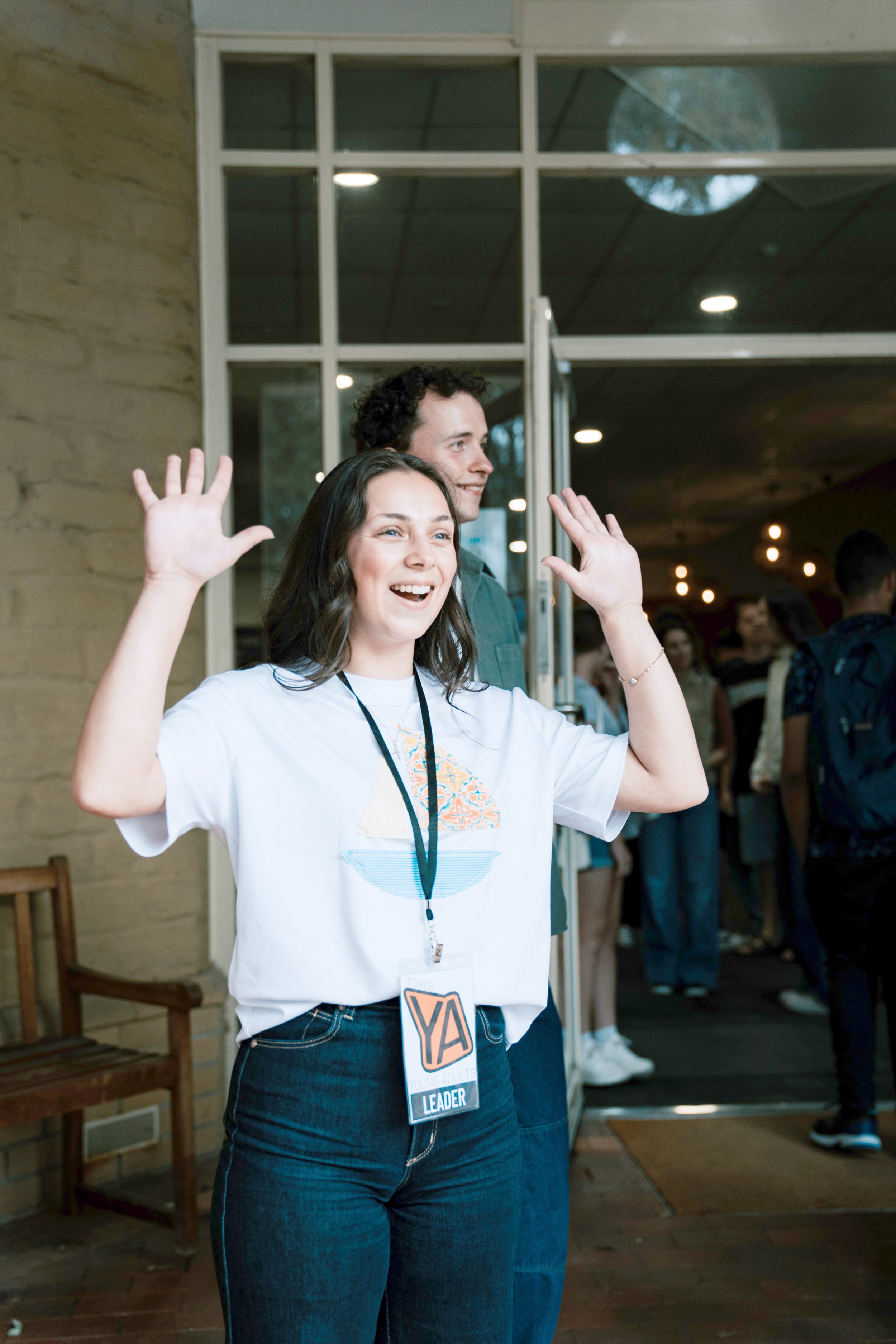 Young woman with long dark hair wearing a white T-shirt with a pizza slice design and a black lanyard with a badge that says 'Youth Leader.' She is smiling and waving with both hands. Behind her, there are people and a young man with curly hair, also smiling. The setting appears to be a gathering or event indoors.