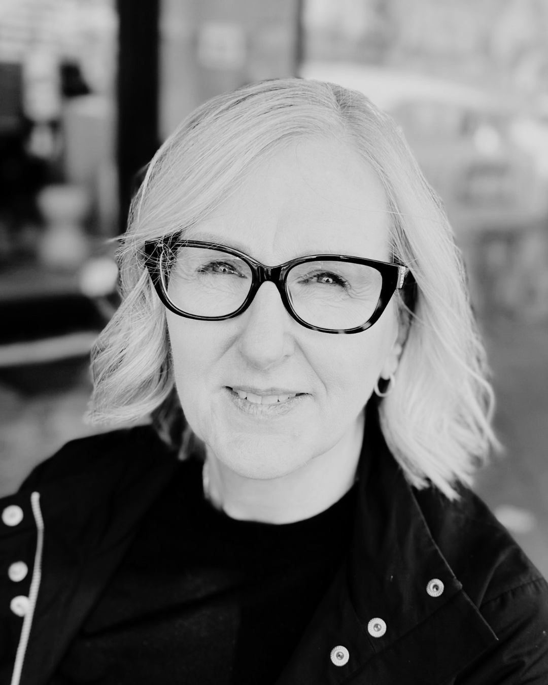 A black and white portrait of a smiling woman with shoulder-length wavy hair wearing glasses and a dark jacket.