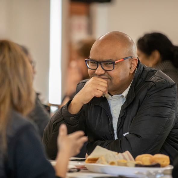 Man in his fifties wearing glasses and a black jacket sitting at a table of food talking to a lady