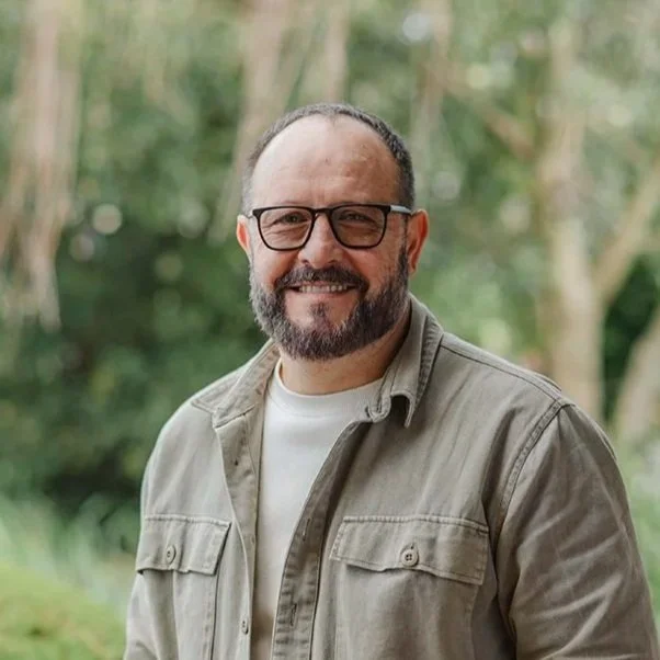 Man in his fifties with a brown beard wearing a beige jacket and white shirt standing in front of bushes outside