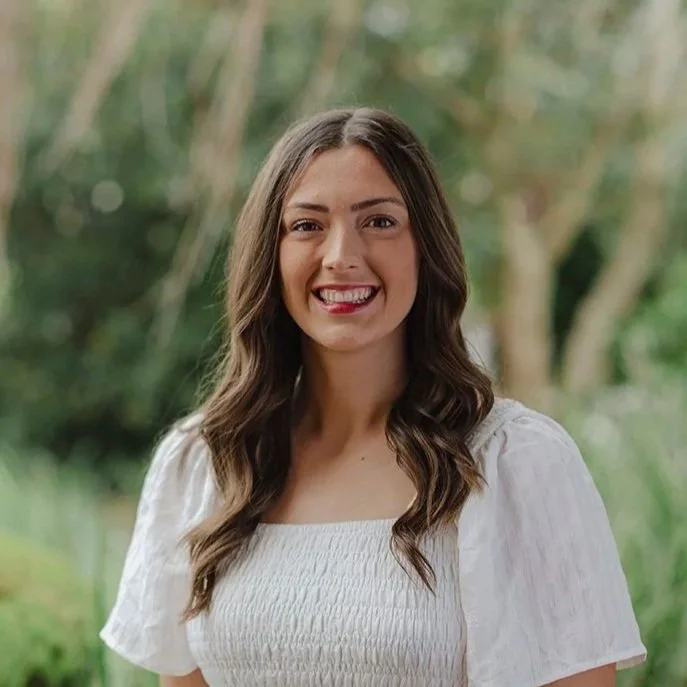 Lady in her twenties wearing a white short length dress standing in front of bushes outside