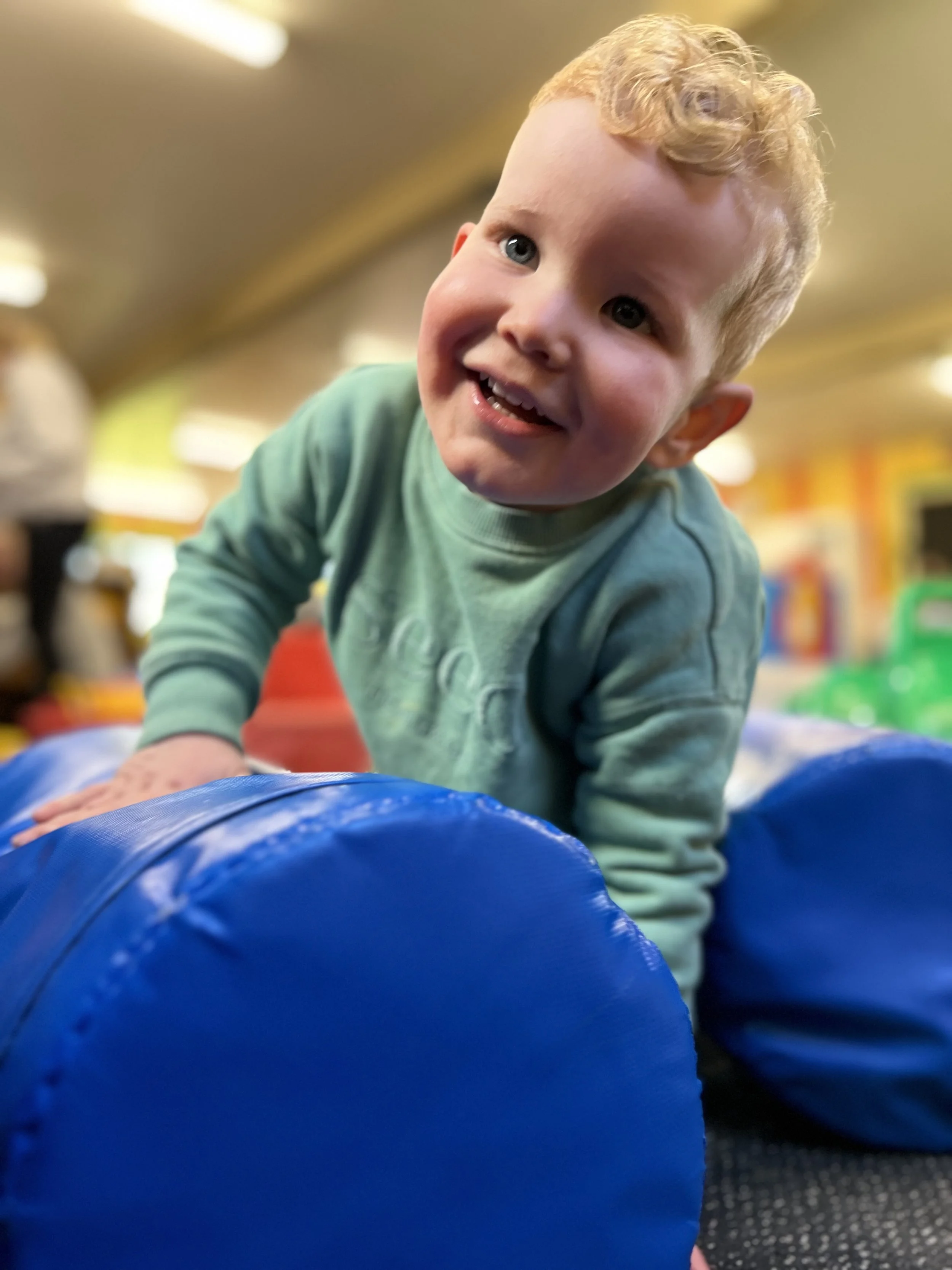 A young boy with blonde curly hair and blue eyes smiling while crawling on a blue padded surface with colorful play equipment in the background.