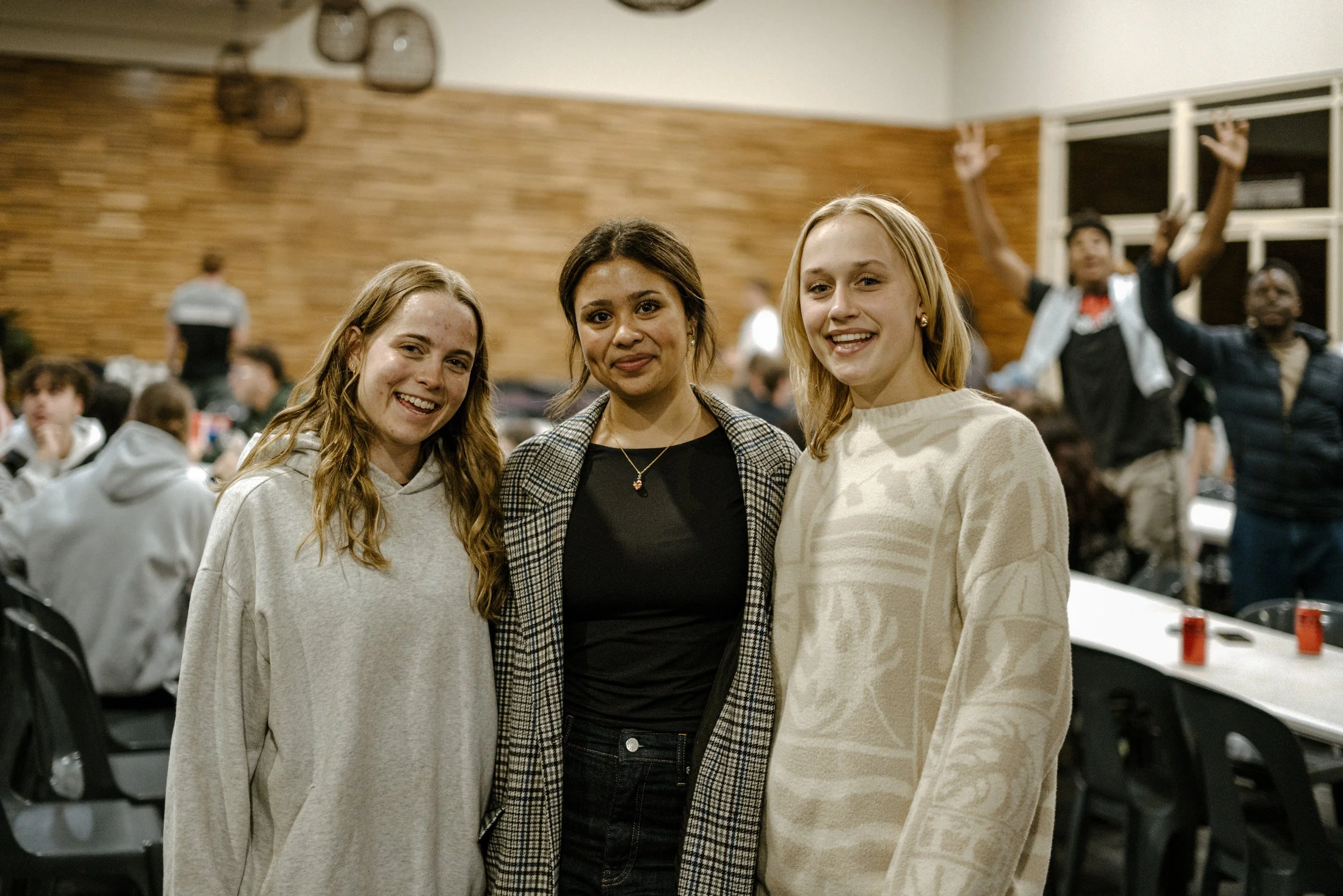 Three young women smiling and posing for a photo in a crowded indoor space, with people in the background.