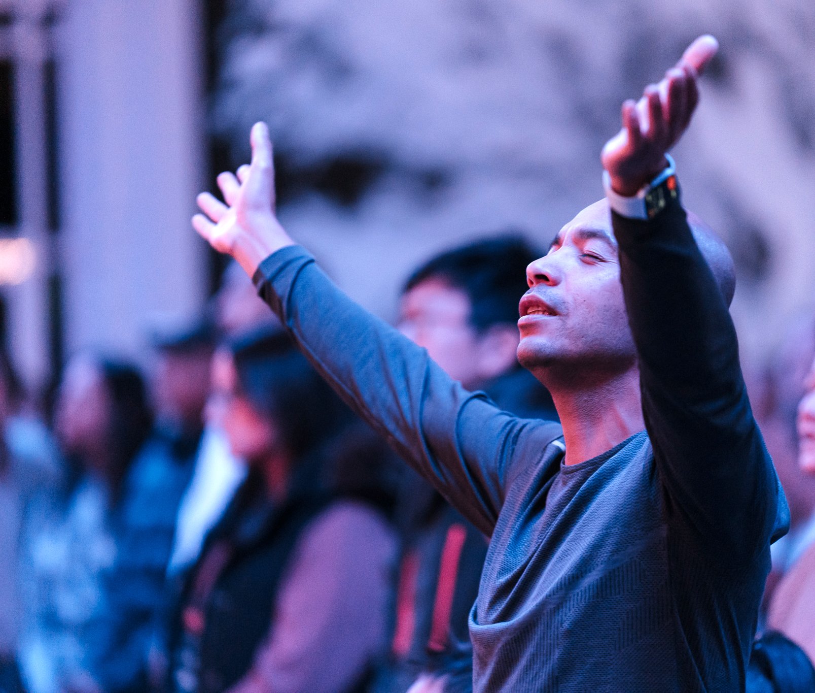Man in his thirties raising both arms while worshiping wearing a blue long sleeve top