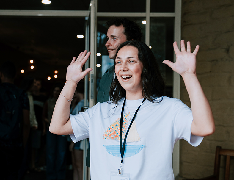 A young woman with dark hair smiling and waving her hands, standing in front of a glass door at an indoor event. There are people and string lights in the background.