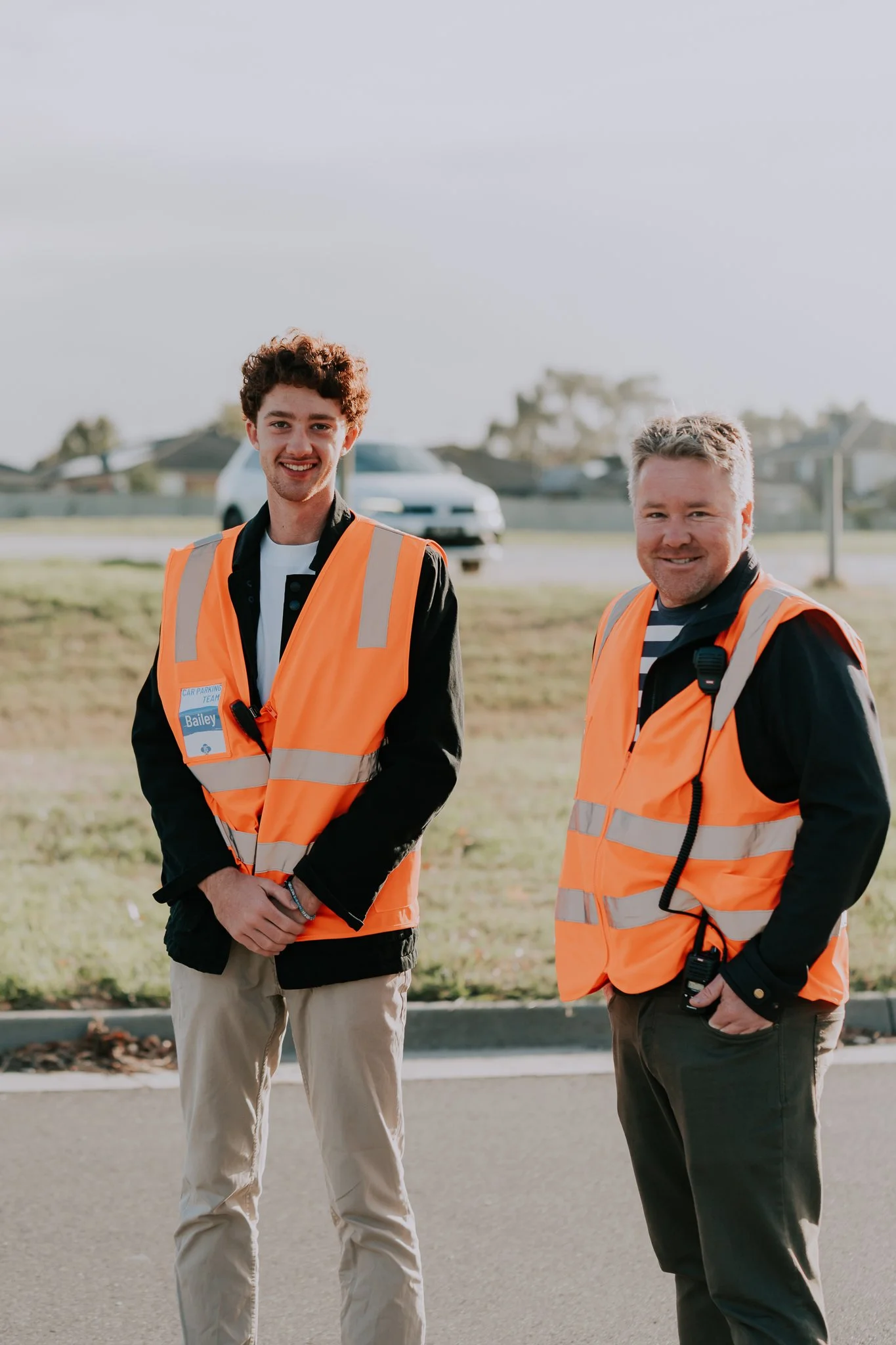Two men wearing orange safety vests standing outdoors on a sidewalk, with a car and houses in the background. The younger man on the left has curly brown hair and is smiling, while the older man on the right has gray hair and is also smiling.