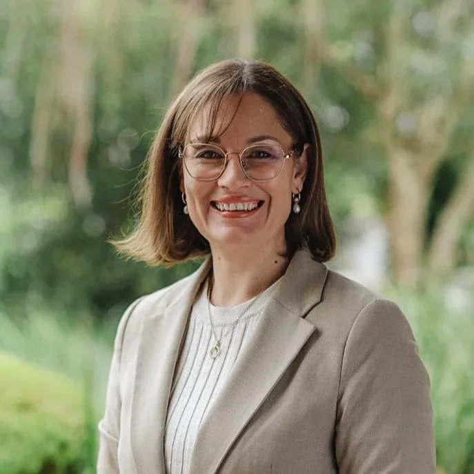 Lady in her thirties wearing glasses and a green top standing in front of bushes outside