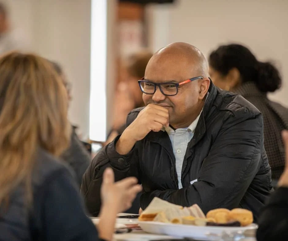 A man in his fifties wearing a black shirt and glasses is talking to a lady at a table
