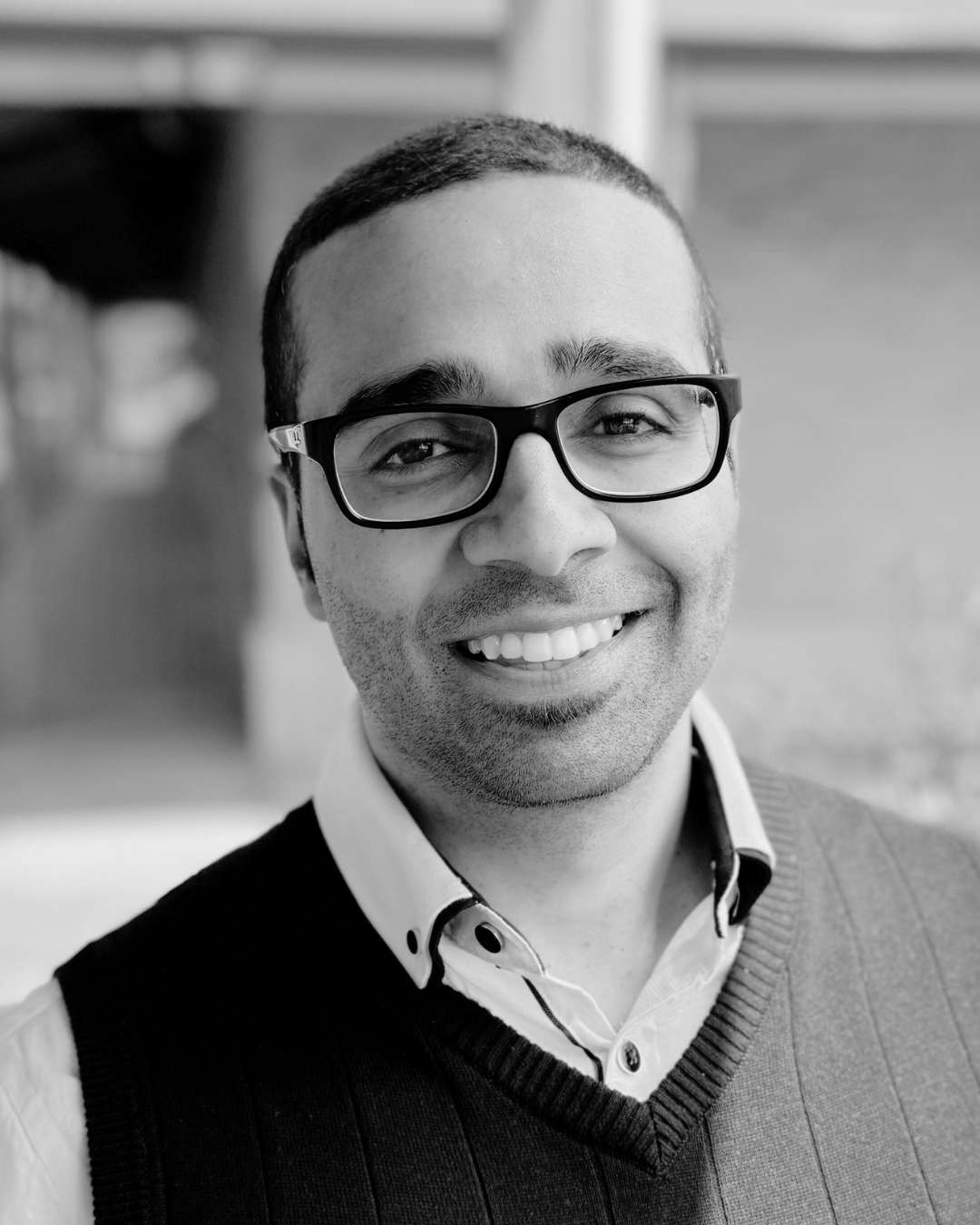 Black and white portrait of a smiling man with short hair, glasses, a collared shirt, and a sweater vest, standing in front of a blurred background.