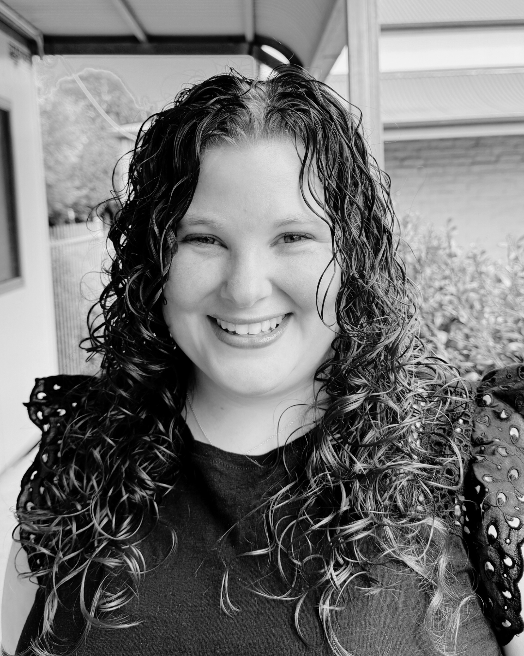 A smiling woman with curly hair, wearing a dark top with eyelet detailing on the sleeves, standing outdoors near a porch or patio area.