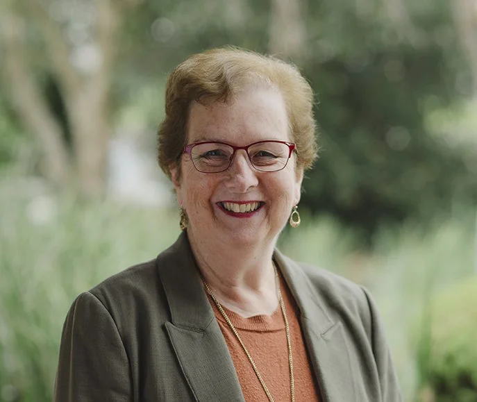 Debbie is wearing glasses and a green blazer and is in her sixties