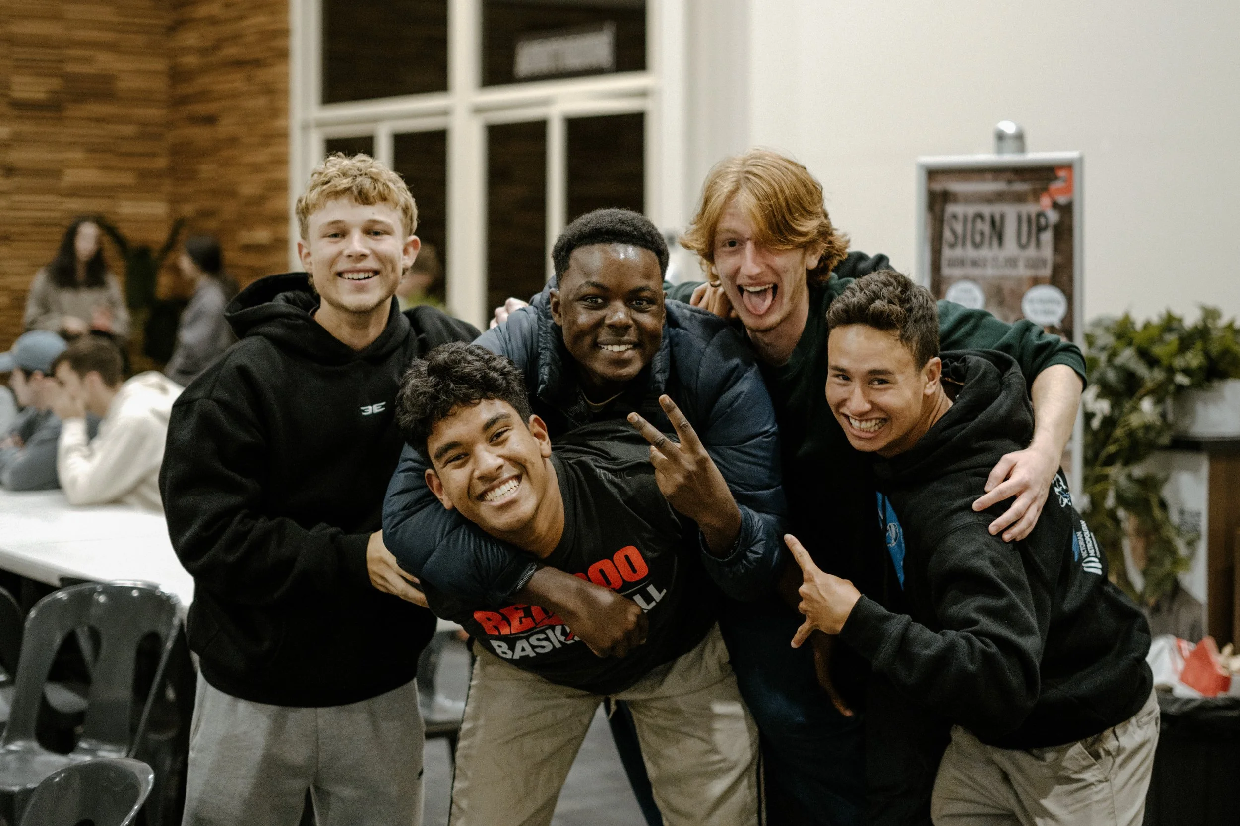 A group of six young men smiling and posing together indoors. They are dressed casually, and some are making hand gestures. In the background, there are other people and a sign that reads 'SIGN UP'.