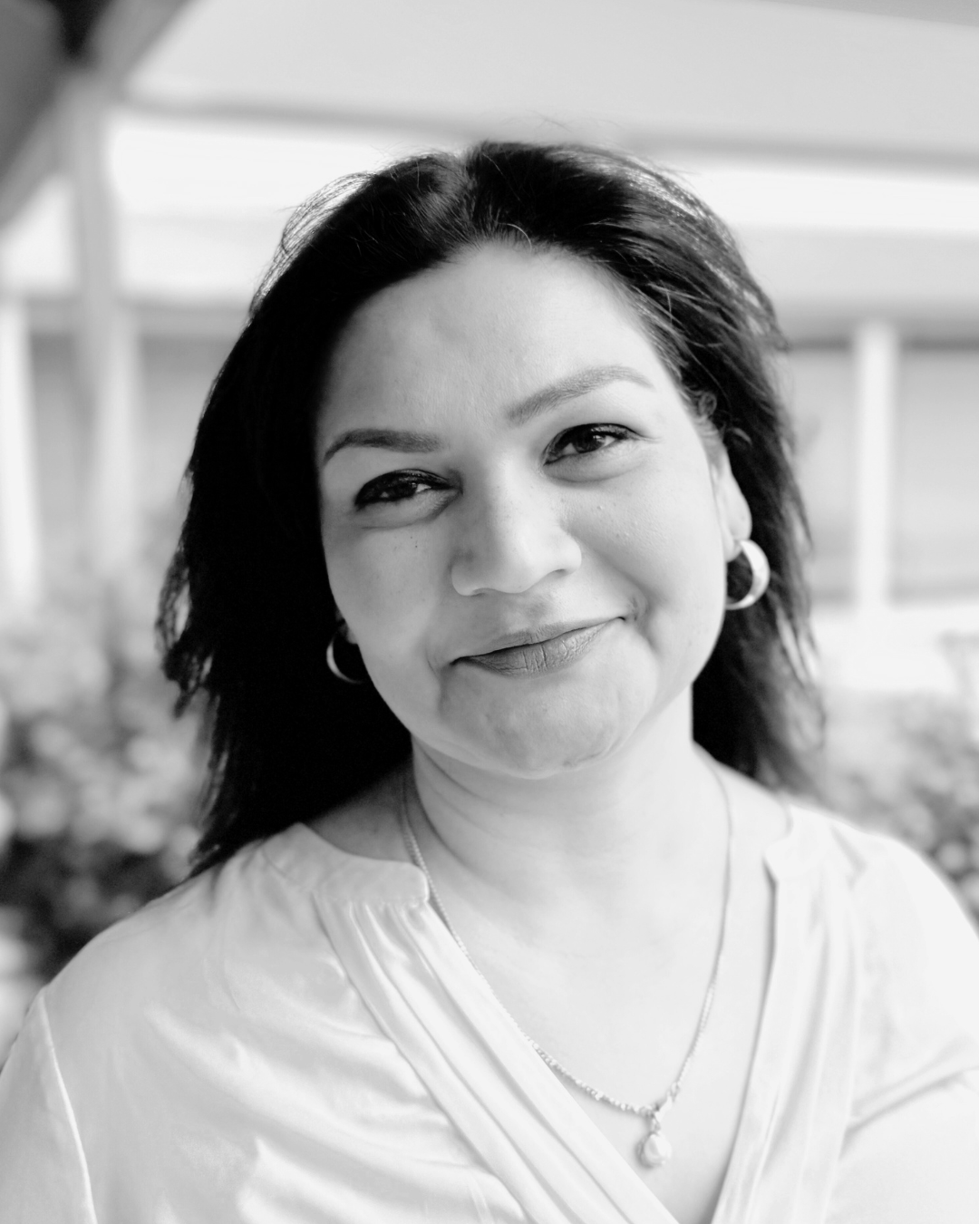 A smiling woman with dark hair and earrings, wearing a light-colored top, in an outdoor setting with blurred background.