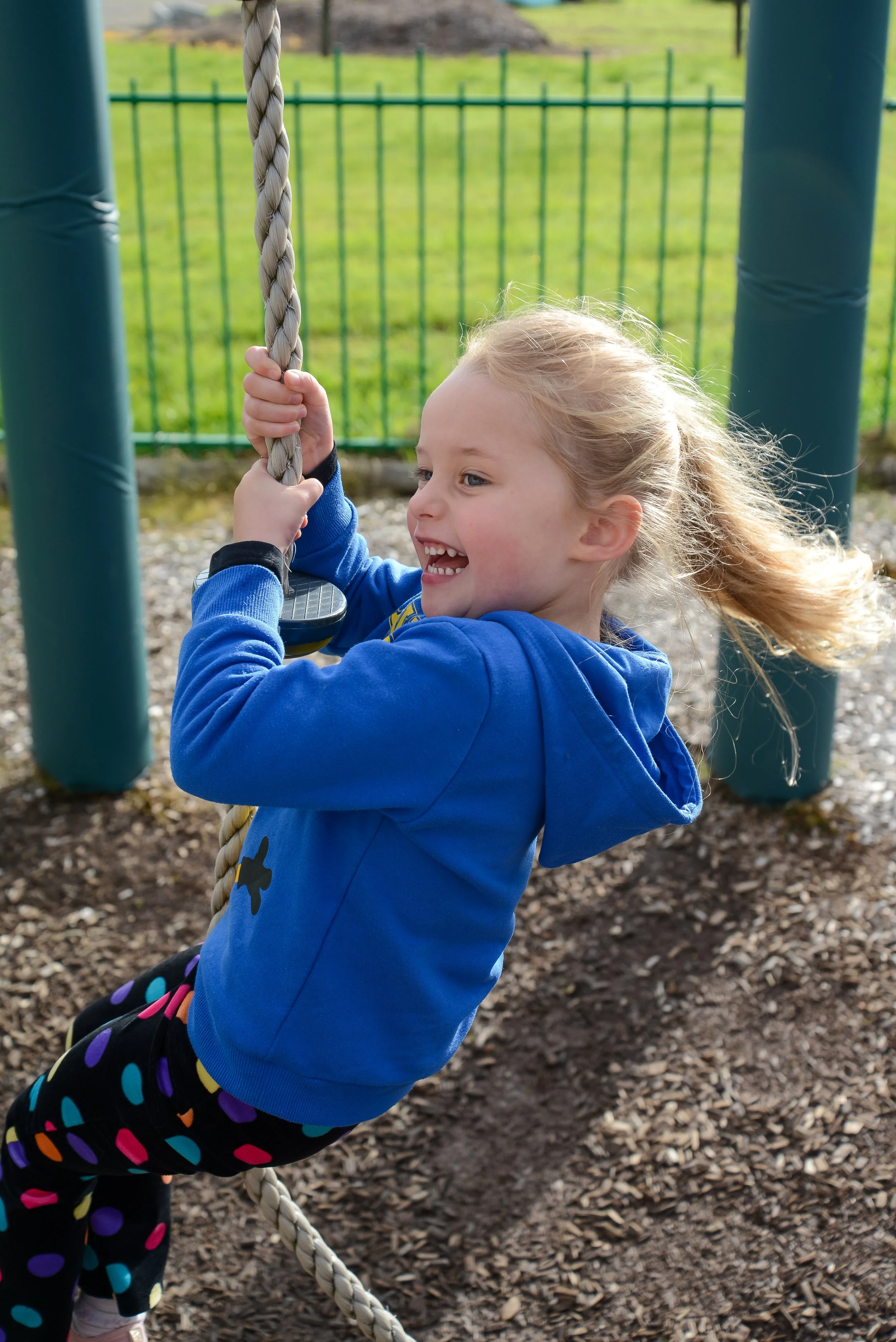 A young girl with blonde hair, wearing a blue hoodie and colorful polka dot pants, enjoying herself on a playground swing set.