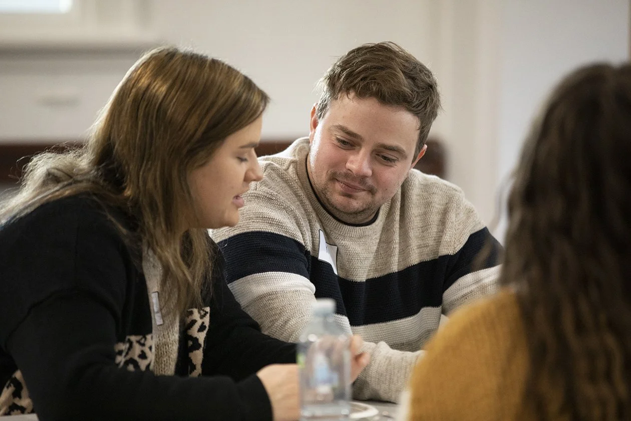 Three people sitting at a table in a meeting or discussion, with one woman speaking and a man listening and smiling.