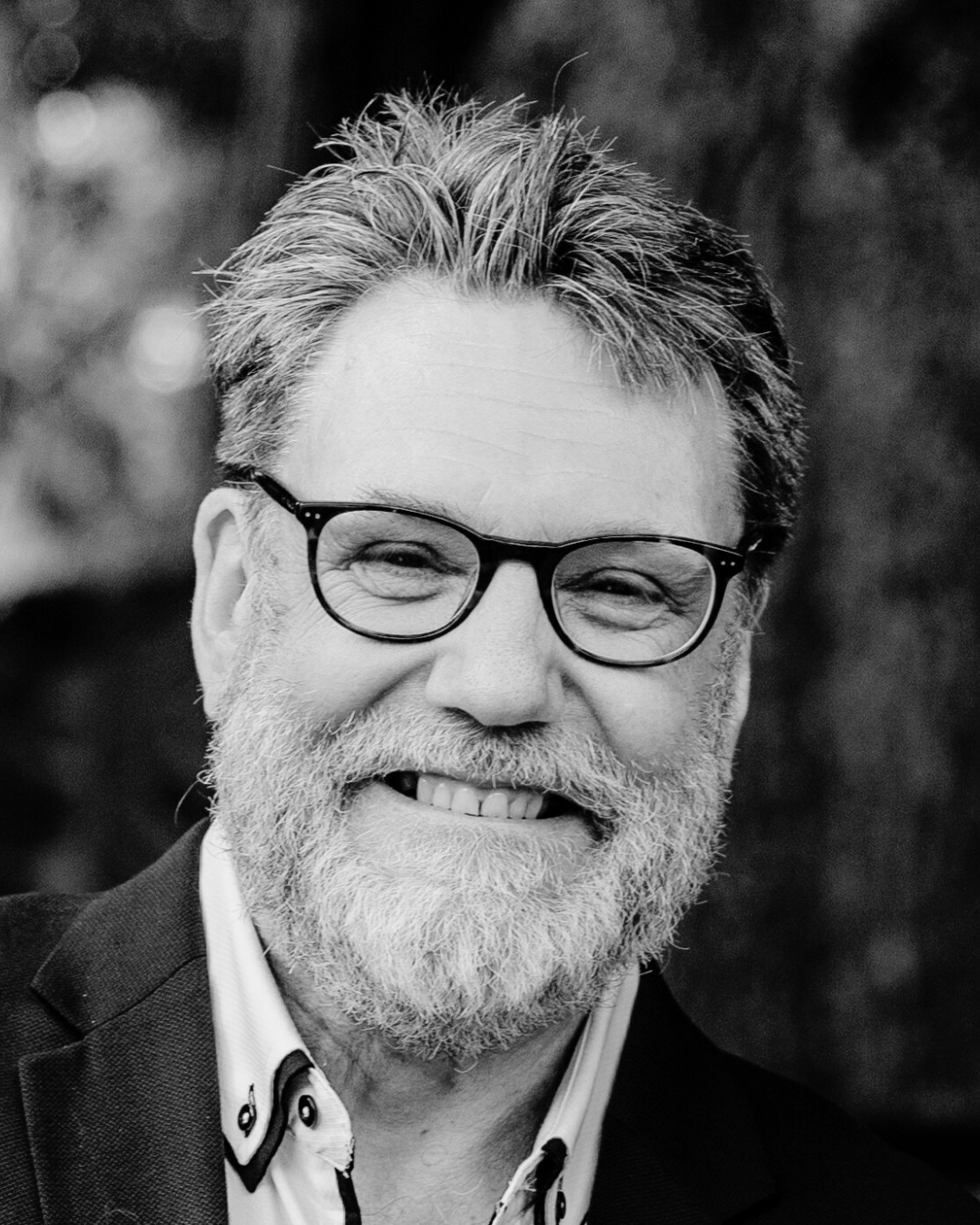A black and white portrait of a smiling older man with glasses, a beard, and styled hair, wearing a suit jacket and dress shirt.