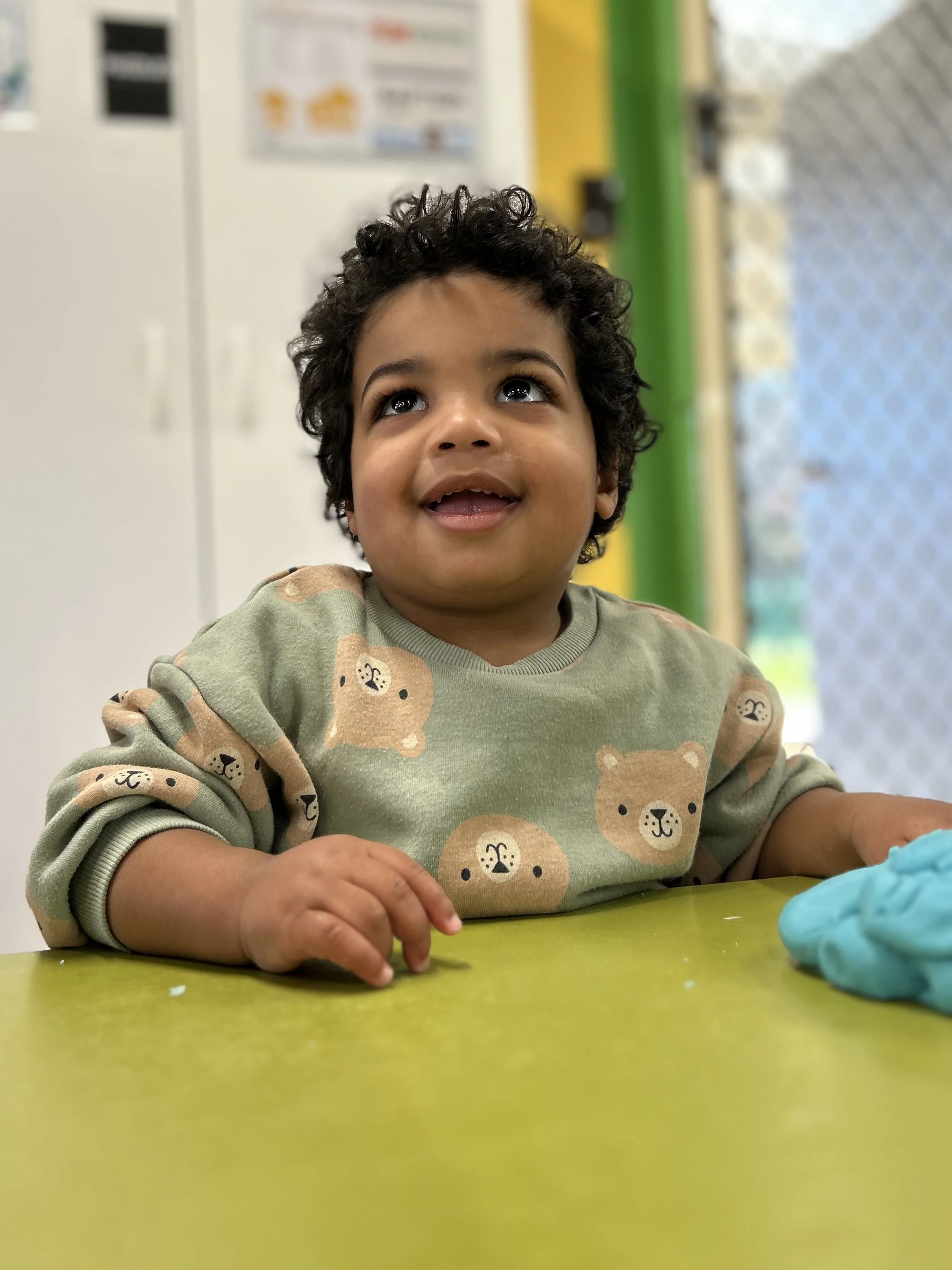 A young child with curly dark hair sitting at a table, looking upward with a happy expression. The child is wearing a beige sweatshirt with a bear face pattern. There are some blue modeling clay or playdough in front of the child on a green table.