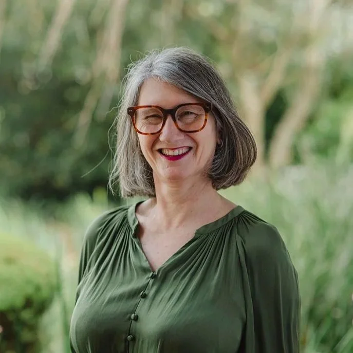 Lady in her fifties wearing a white dress wearing red glasses standing in front of bushes outside