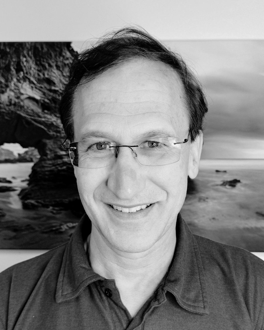 A man with glasses and a collared shirt smiling, standing in front of a large rock formation on a beach, with the ocean in the background.