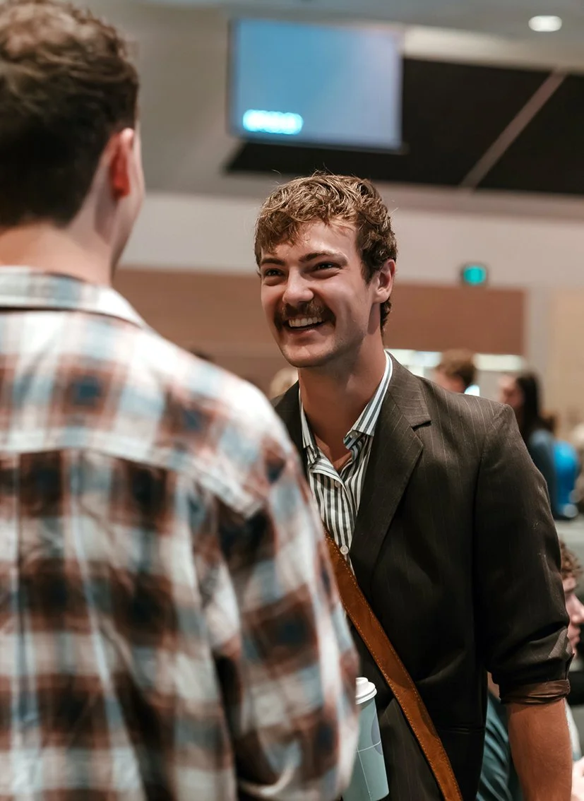Young man in his twenties wearing a brown jacket talking to a friend