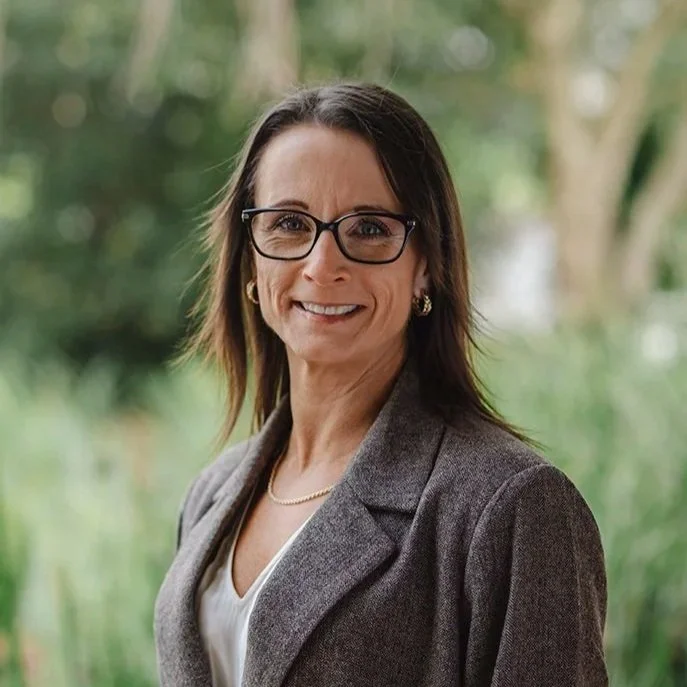 A lady in her forties wearing glasses and a beige shirt standing in front of bushes outside