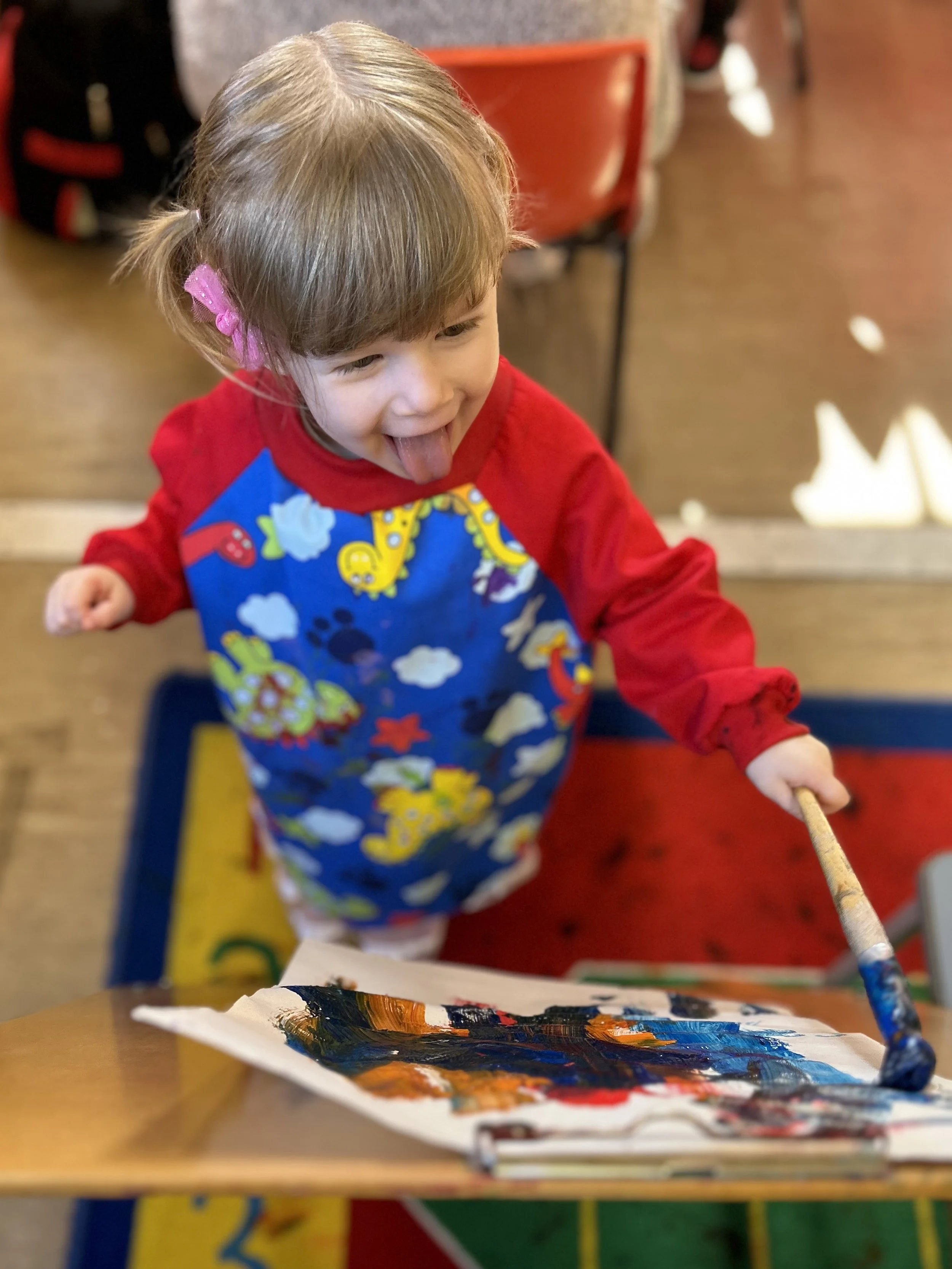 A young child with light brown hair, wearing a pink hair clip and colorful apron, is smiling and sticking out their tongue while painting with a brush on paper.
