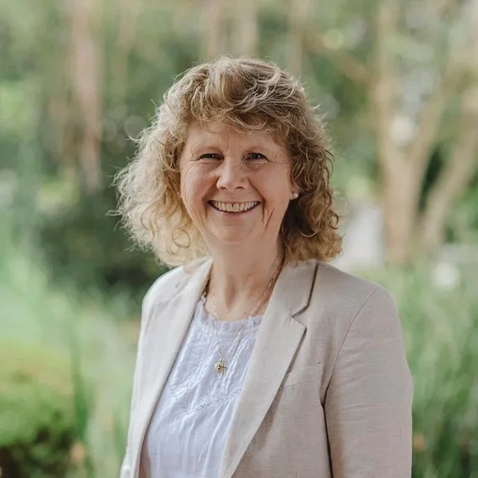 Lady in her sixties wearing a white t-shirt with beige jacket standing in front of bushes outside