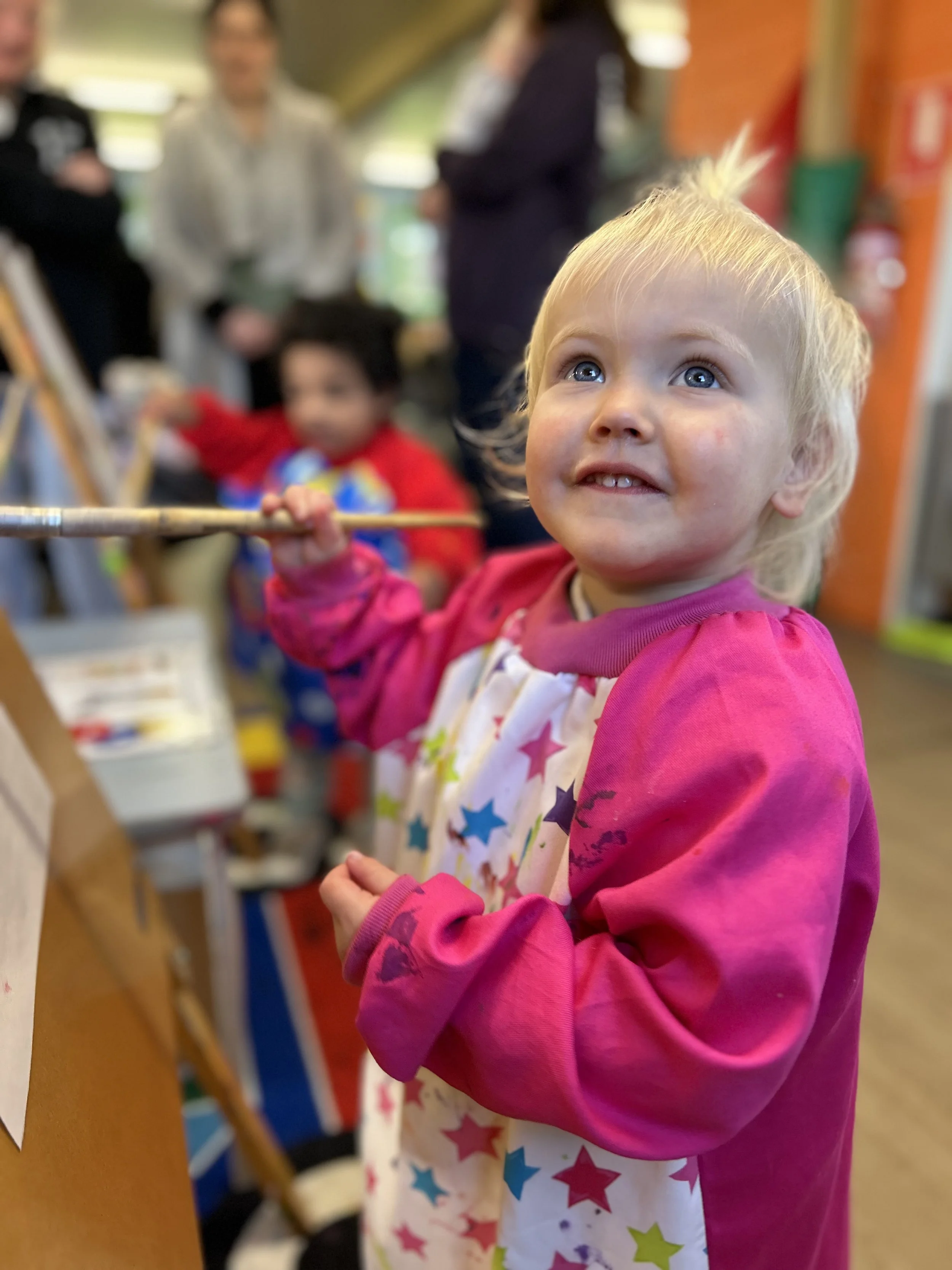 A young blonde girl with blue eyes and freckles, wearing a pink long-sleeve shirt and a star-patterned apron, is looking upward while holding a paintbrush in her right hand in an art classroom. In the blurred background, other children and adults are engaged, and there are colorful walls and shelves.