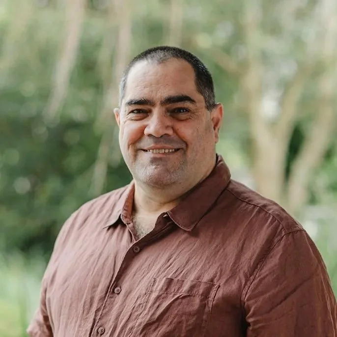 Man in his fifties clean shaven wearing a beige short sleeve shirt standing in front of bushes outside