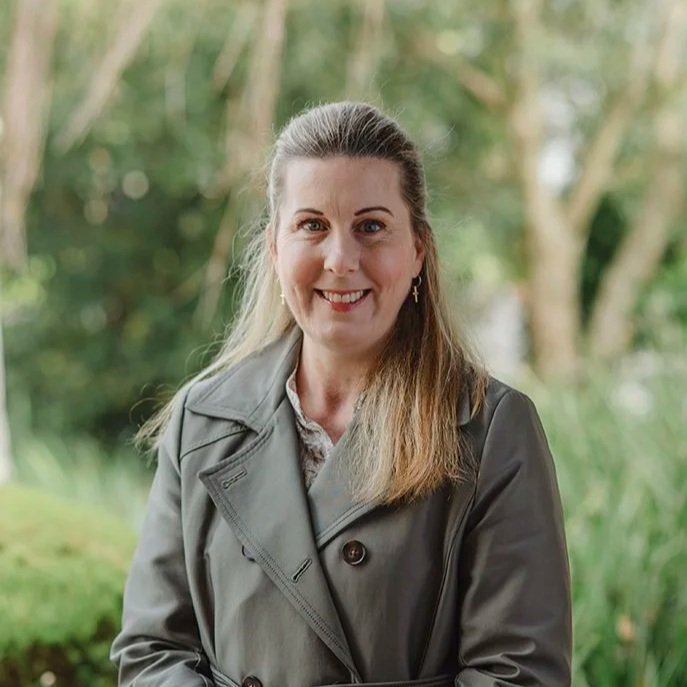 A lady in her fifties wearing a green jacket standing in front of bushes outside