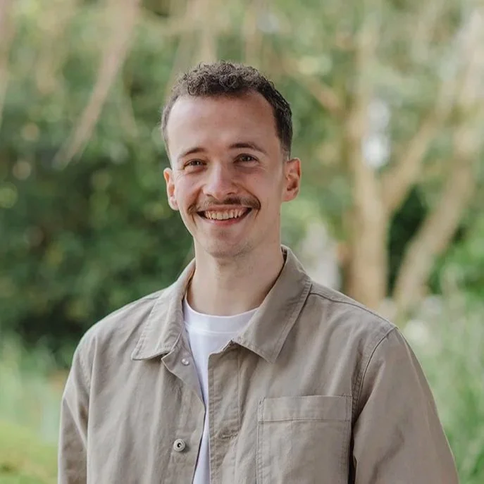 Man in his twenties wearing a green shirt standing in front of bushes outside