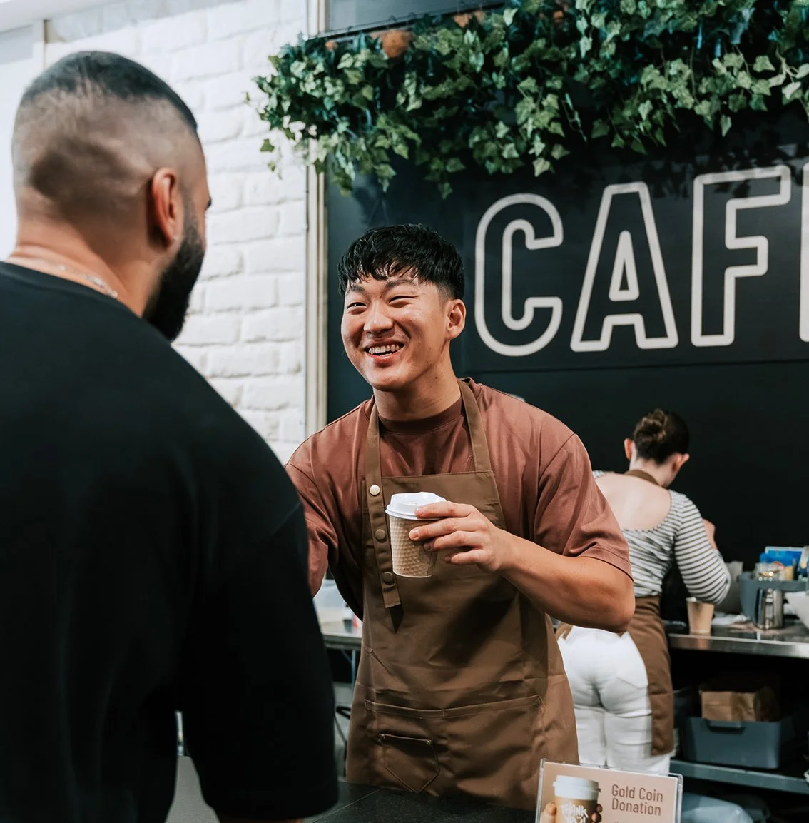Young man making a coffee wearing an apron and handing a take away coffee to a visitor