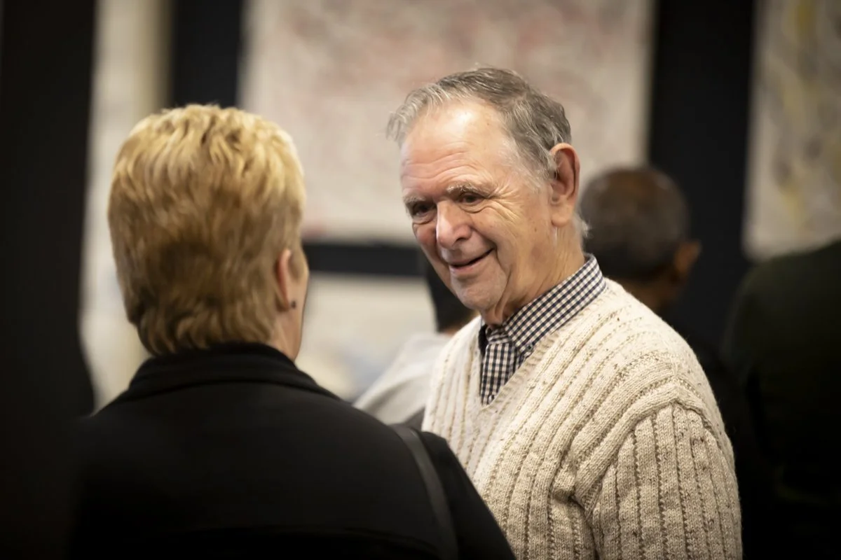 Two older people having a conversation at an event. The man is smiling and wearing a sweater over a button-up shirt, while the woman has short blonde hair and wears a dark top.