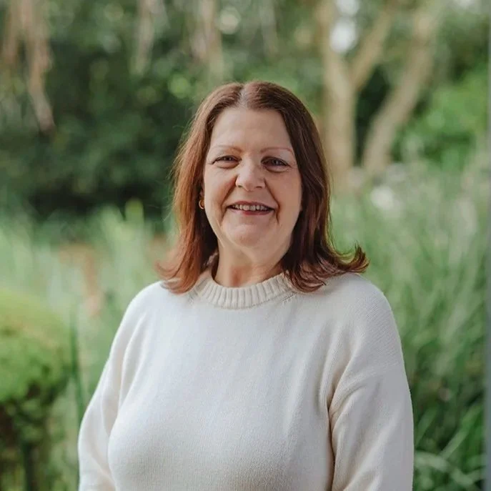 A lady in her sixties wearing a pink shirt standing in front of bushes outside
