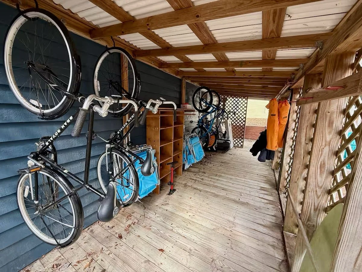 A covered outdoor storage area with bicycles hanging on the wall, a wooden shelf, and jackets hanging on hooks.