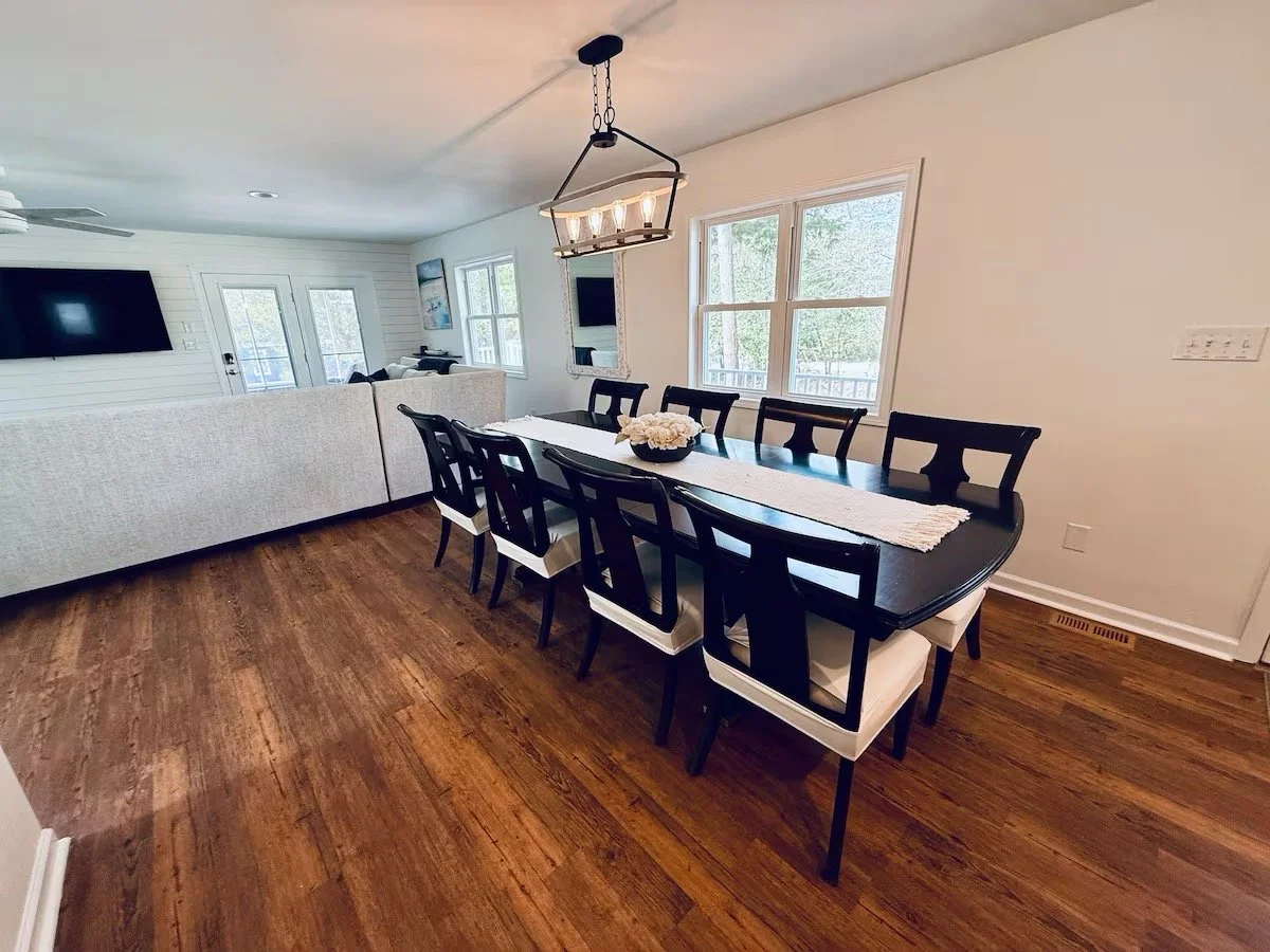 Dining room with a black table, eight matching chairs, a white table runner, a flower centerpiece, hardwood floors, windows, and a light fixture hanging above the table.