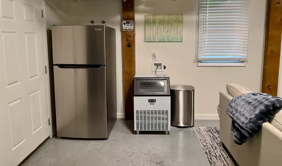 Kitchen corner with a silver refrigerator, a stainless steel ice machine, a metallic trash can, a window with blinds, and a beige sofa with a gray blanket draped over the armrest.