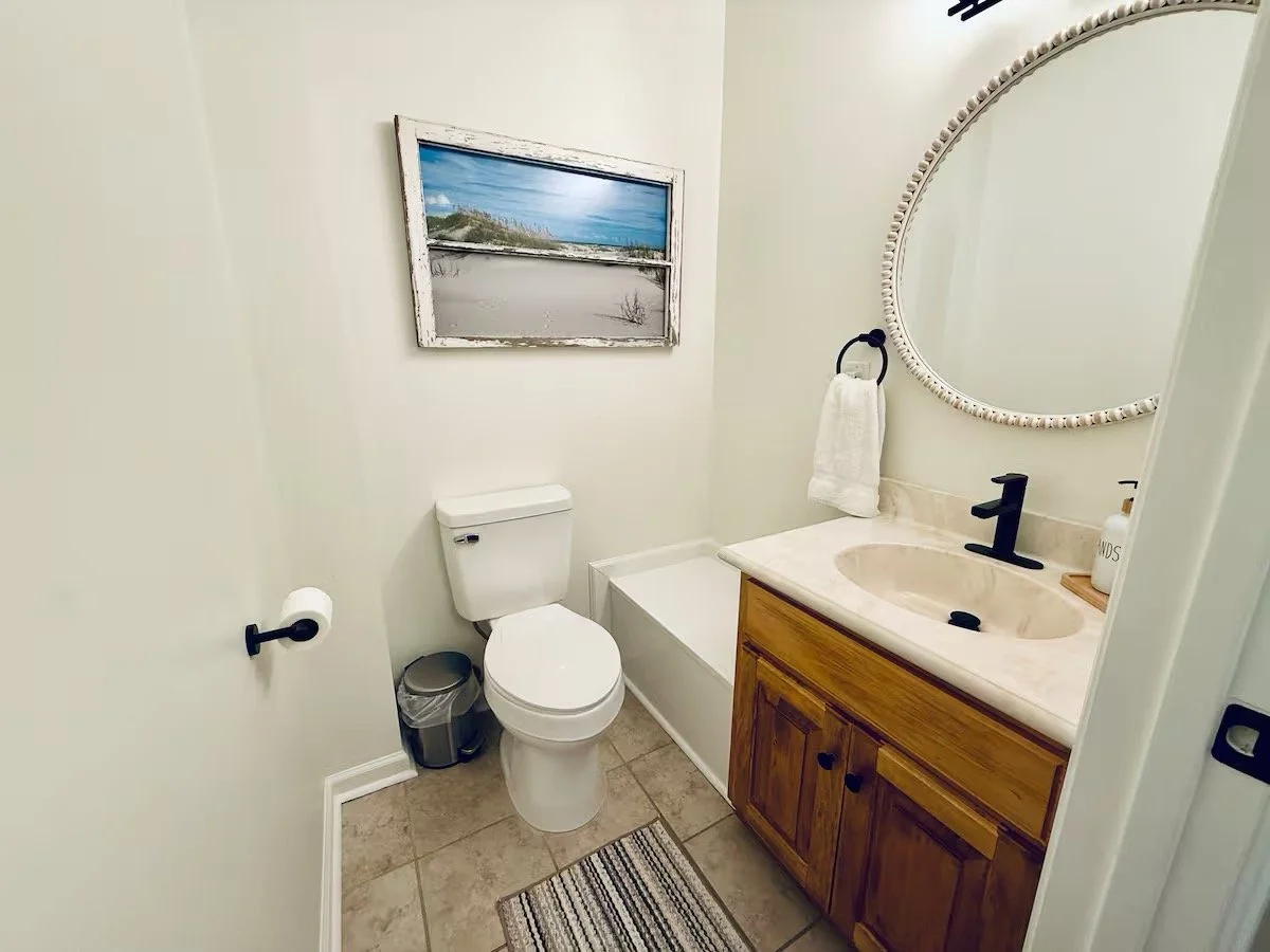 Small bathroom with a toilet, a wooden vanity with a sink and black faucet, a round mirror with a beaded frame, a towel hanging on a ring, a framed beach landscape hanging on the wall, a bathtub, a trash can, and a striped rug on tiled floor.