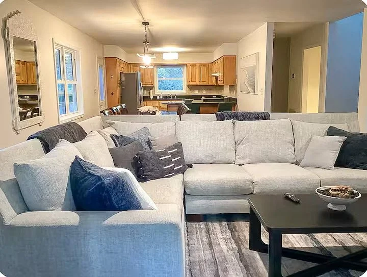 Living room with a large white sectional sofa, various pillows, a black coffee table with a remote and a bowl of snacks, in front of a kitchen area with wooden cabinets and a stainless steel refrigerator.