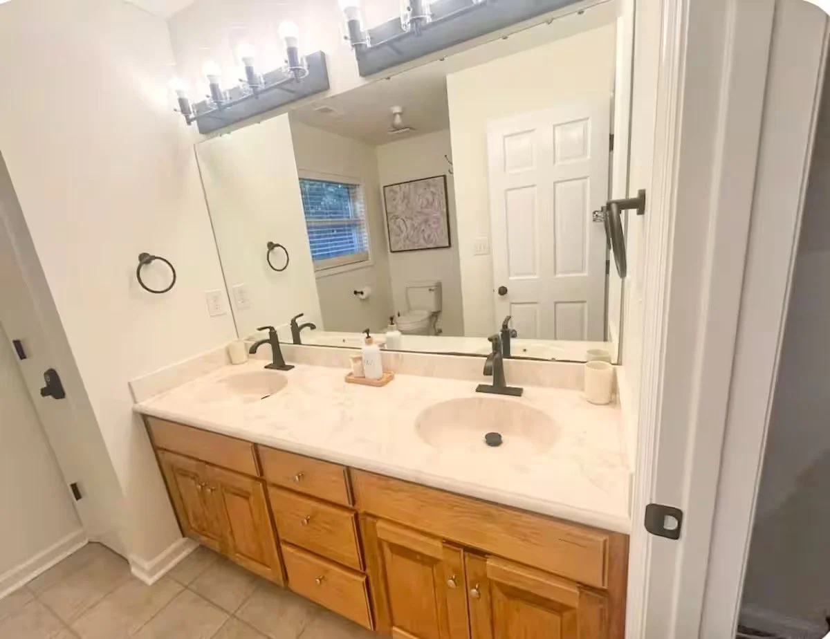 Bathroom with double vanity sink, large mirror, black fixtures, wood cabinets, wall-mounted light fixture, and a toilet in the background.