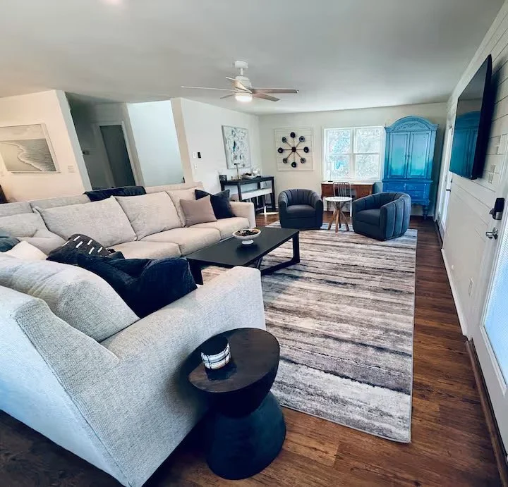 A modern living room with a white sofa, black pillows, black coffee table, a black side table with a candle, two black armchairs, a blue armoire, a striped area rug, a ceiling fan, and large windows with white walls and wood flooring.