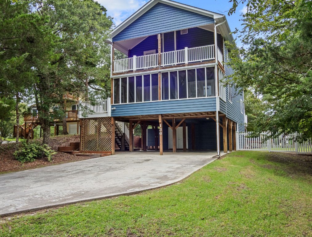 A blue two-story house on stilts with a screened porch and balcony, surrounded by green trees and a concrete driveway.
