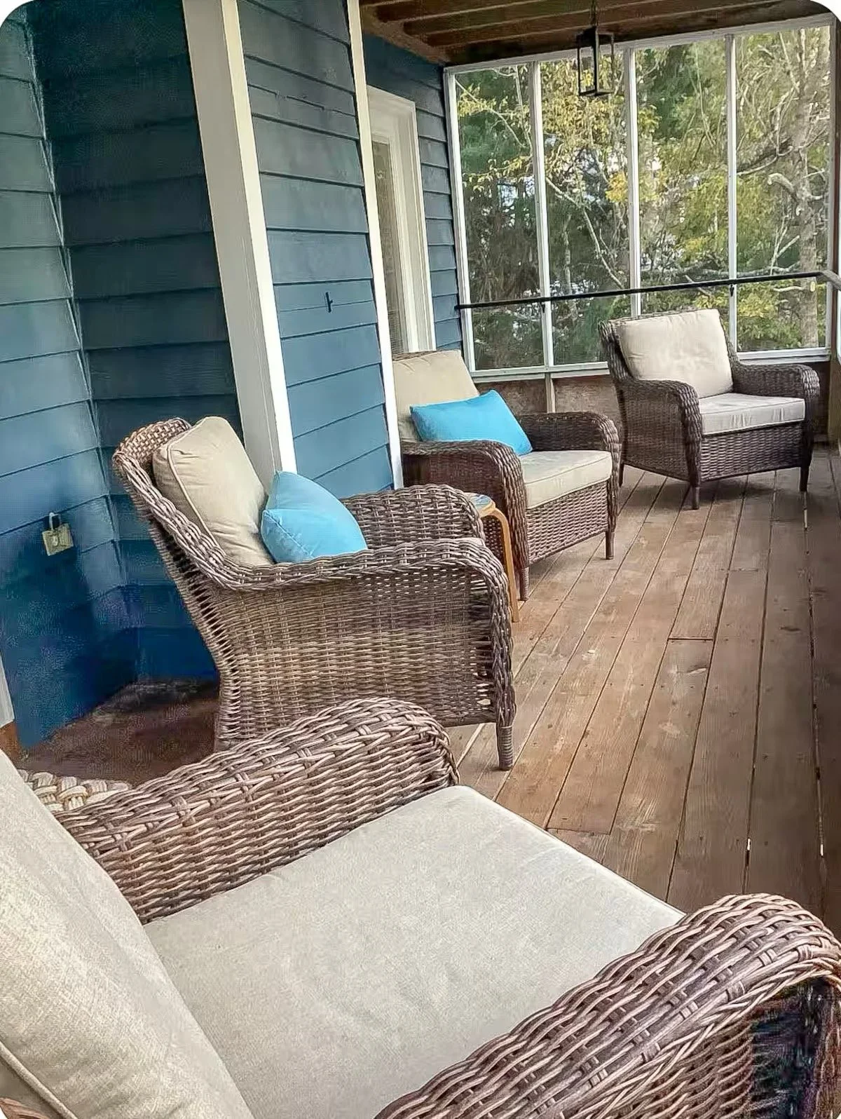 A screened porch with wicker and cushioned chairs, some with blue pillows, and a wooden floor, overlooking trees.